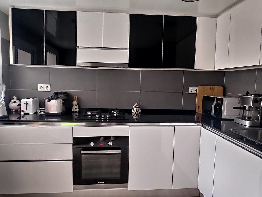 una cocina con muebles blancos, horno y mármoles negros, paredes grises.
