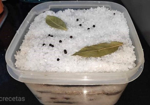 container full of anchovies and coarse salt, on top black peppercorns and a bay leaf