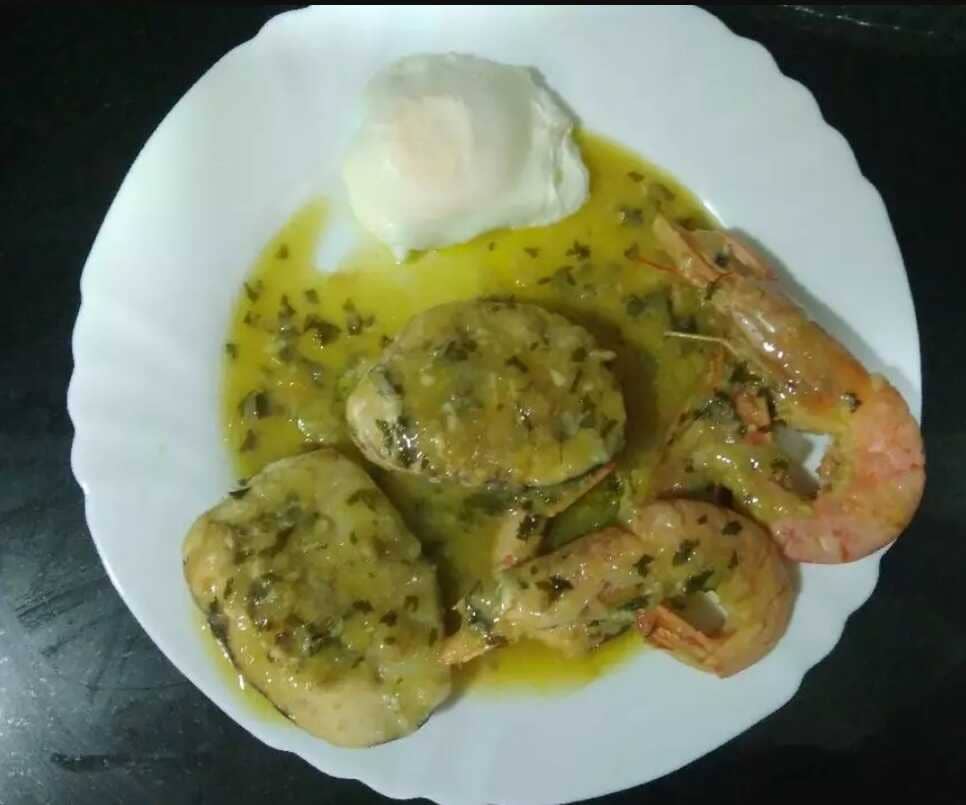 En un plato blanco dos rodajas de merluza, dos gambas y un huevo pochado regado de salsa con perejil.